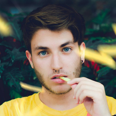 A young man with blue eyes, a septum piercing, and light stubble eating a french fry against a leafy green background.
