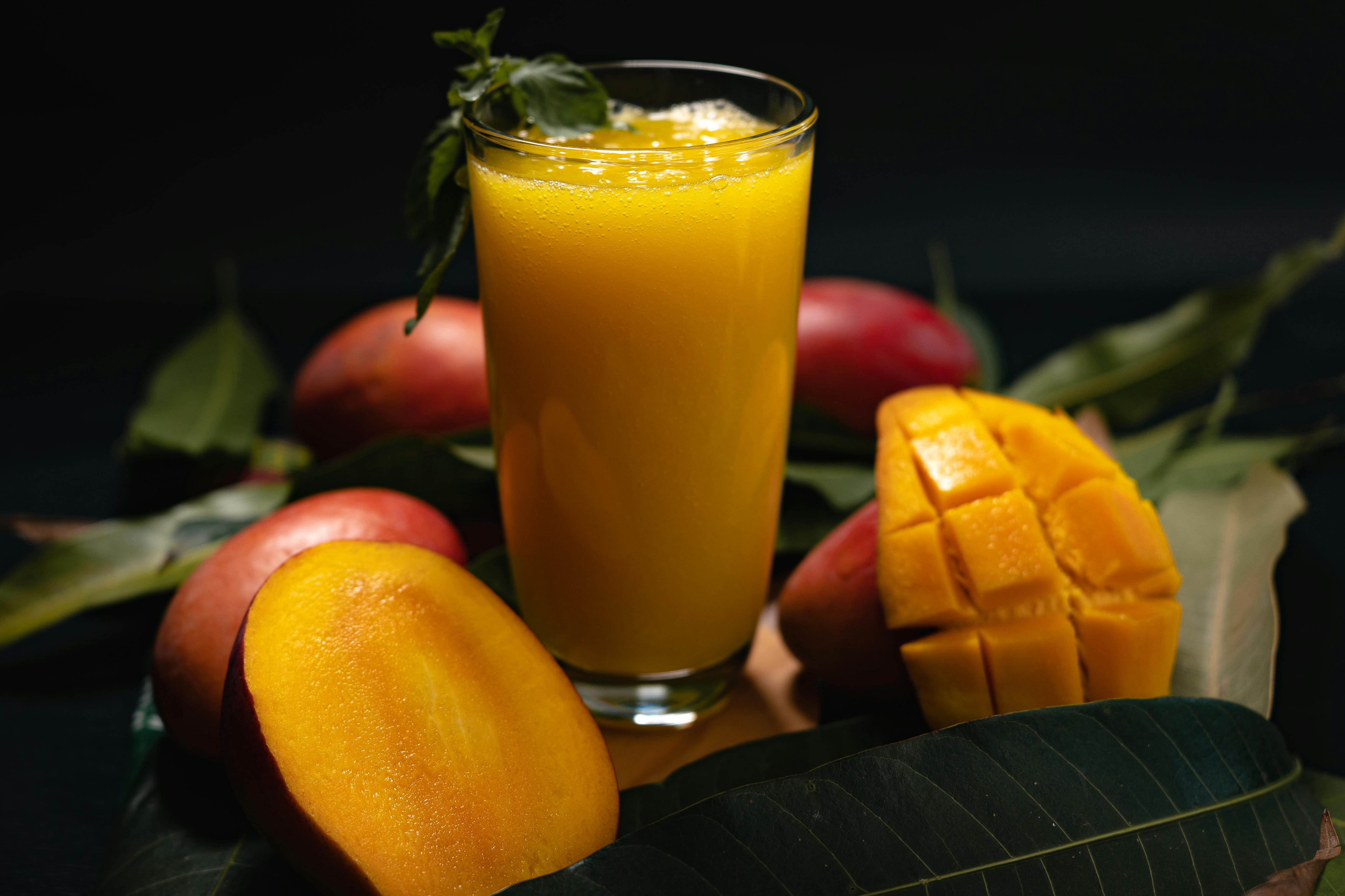 Glass of fresh mango juice with whole and sliced mangoes on leaves.